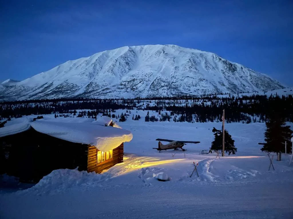Snowbound log cabin and bush plane at twilight beneath an Alaska peak